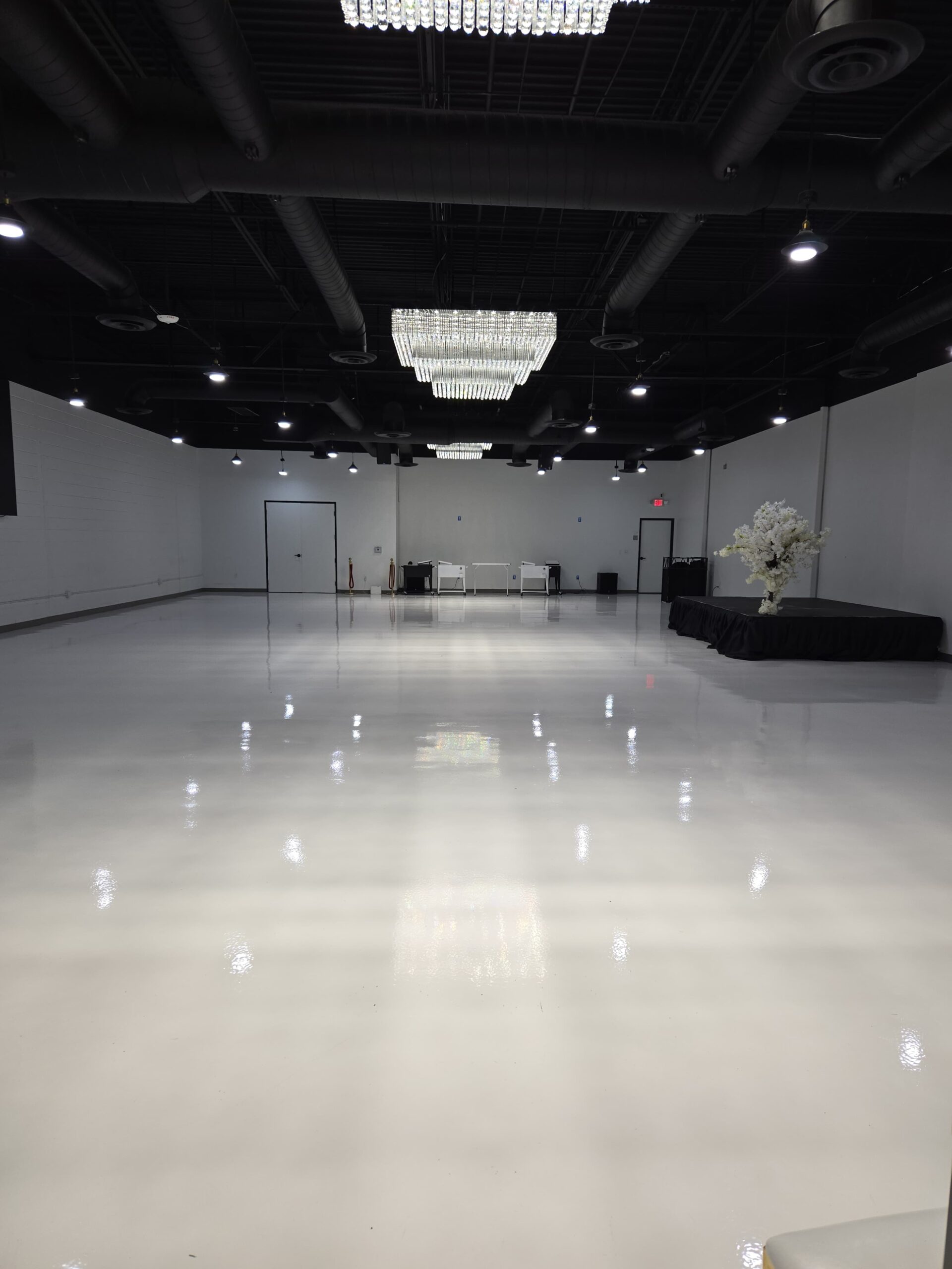 Step into a space where sophistication meets versatility. This image captures our pristine event hall, featuring a high-gloss white floor that reflects the glow of our signature chandeliers and ambient lighting. With a spacious layout, modern black ceiling, and a raised platform perfect for performances or presentations, The Ace Event Venue is designed to transform seamlessly for weddings, corporate gatherings, birthday celebrations, and more.